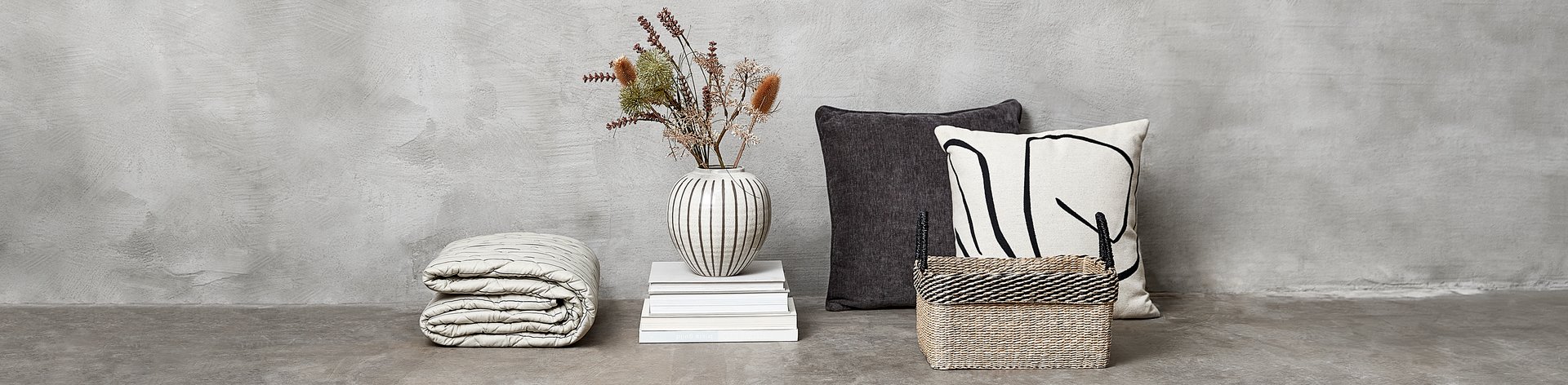 Wohnaccessoires stehen vor einer Betonwand. Darunter eine Vase mit Blumen, ein Korb sowie zwei Kissen
