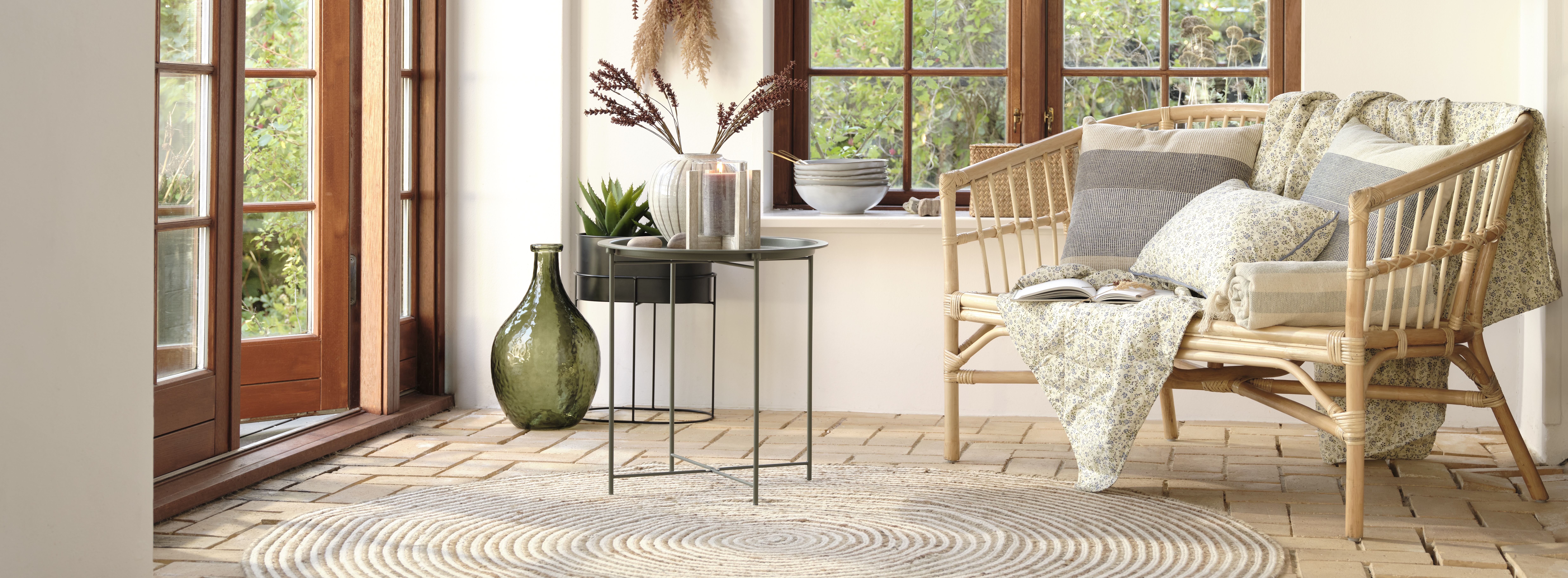 Eine Bank in Bambusoptik mit Kissen und Decken sowie ein Beistelltisch mit einer Vase und Kerze. Im Hintergrund sieht man eine Fensterfront sowei weitere Wohnaccessoires