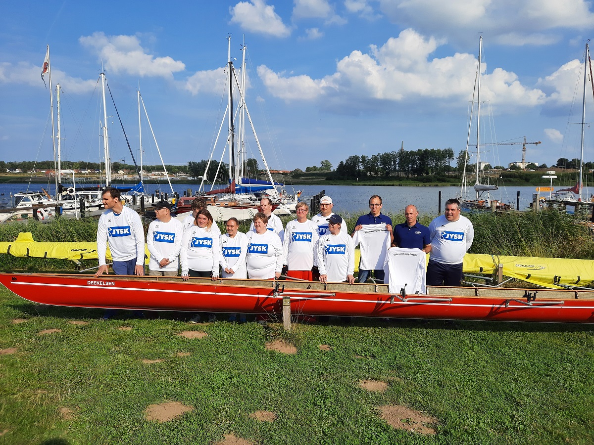 Mehrere Menschen stehen hinter einem Ruderboot. Sie tragen neue Trikots