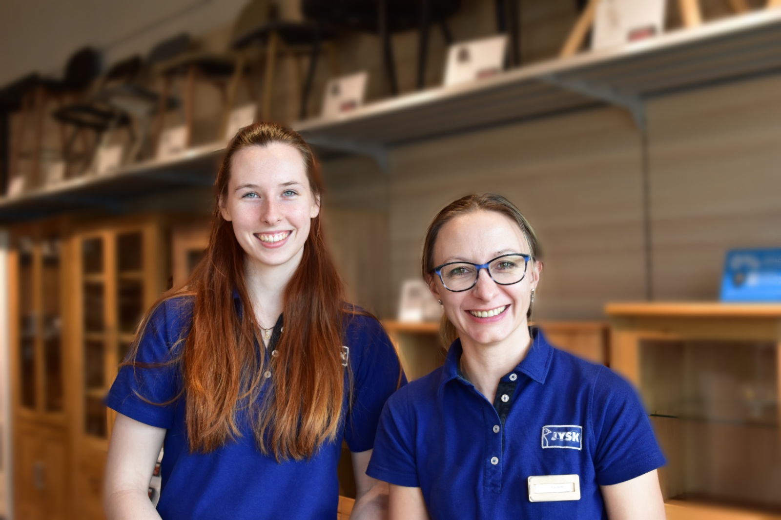 Zwei Frauen – eine mit langen Haaren und eine mit Pferdeschwanz und Brille – lächeln in die Kamera. Beide tragen JYSK-Poloshirts 