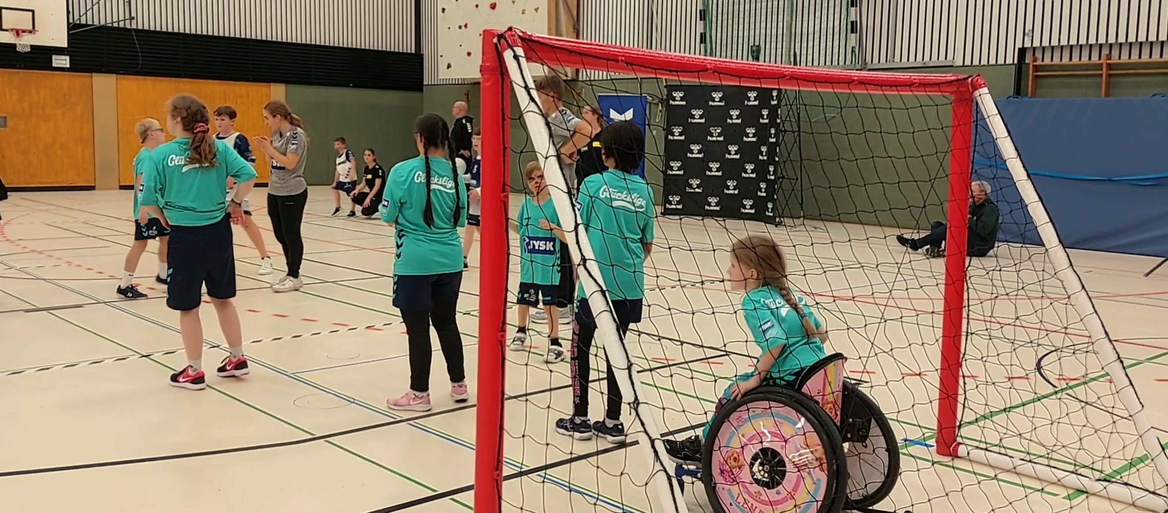 Mehrere Kinder stehen auf einem Handballfeld, im Tor sitzt ein kleines Mädchen mit Pferdeschwanz in einem Rollstuhl und verfolgt konzentriert das Spiel