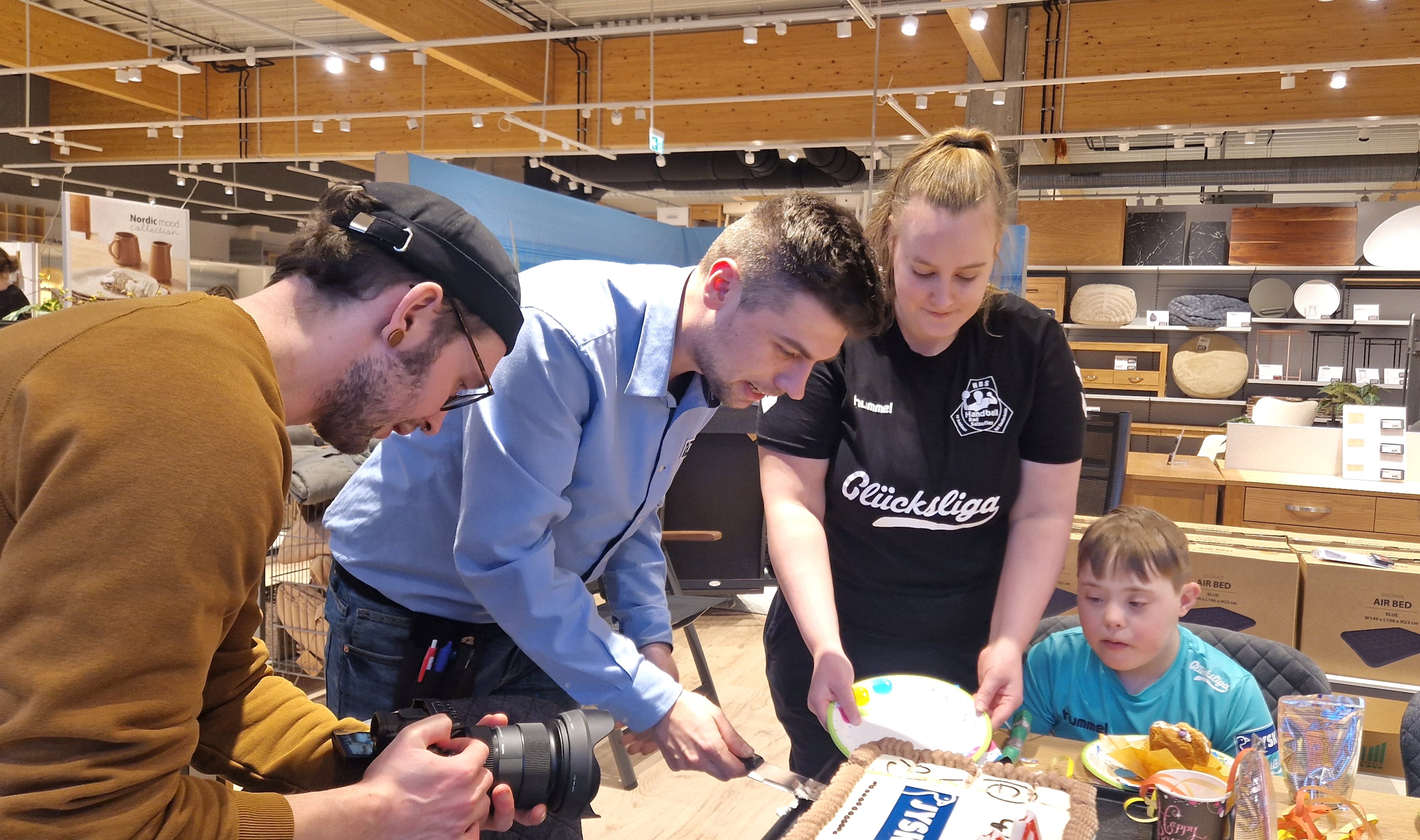 Ein Mann schneidet eine Torte an. Eine Frau steht daneben und hält einen Teller. Ein Mann hält eine Kamera in der Hand und filmt die Szene.