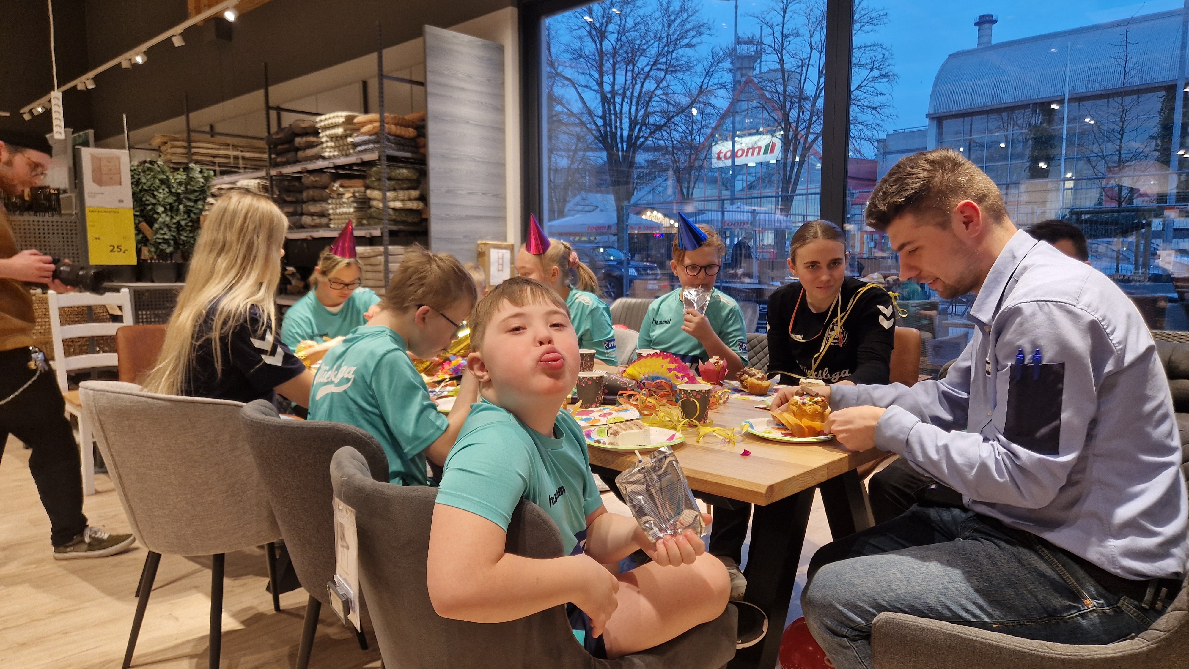 Ein Junge schaut in die Kamera. Er streckt die Zunge heraus. Viele weitere Kinder sind hinter ihm zu sehen. Sie sitzen an einem Tisch und Essen Kuchen.