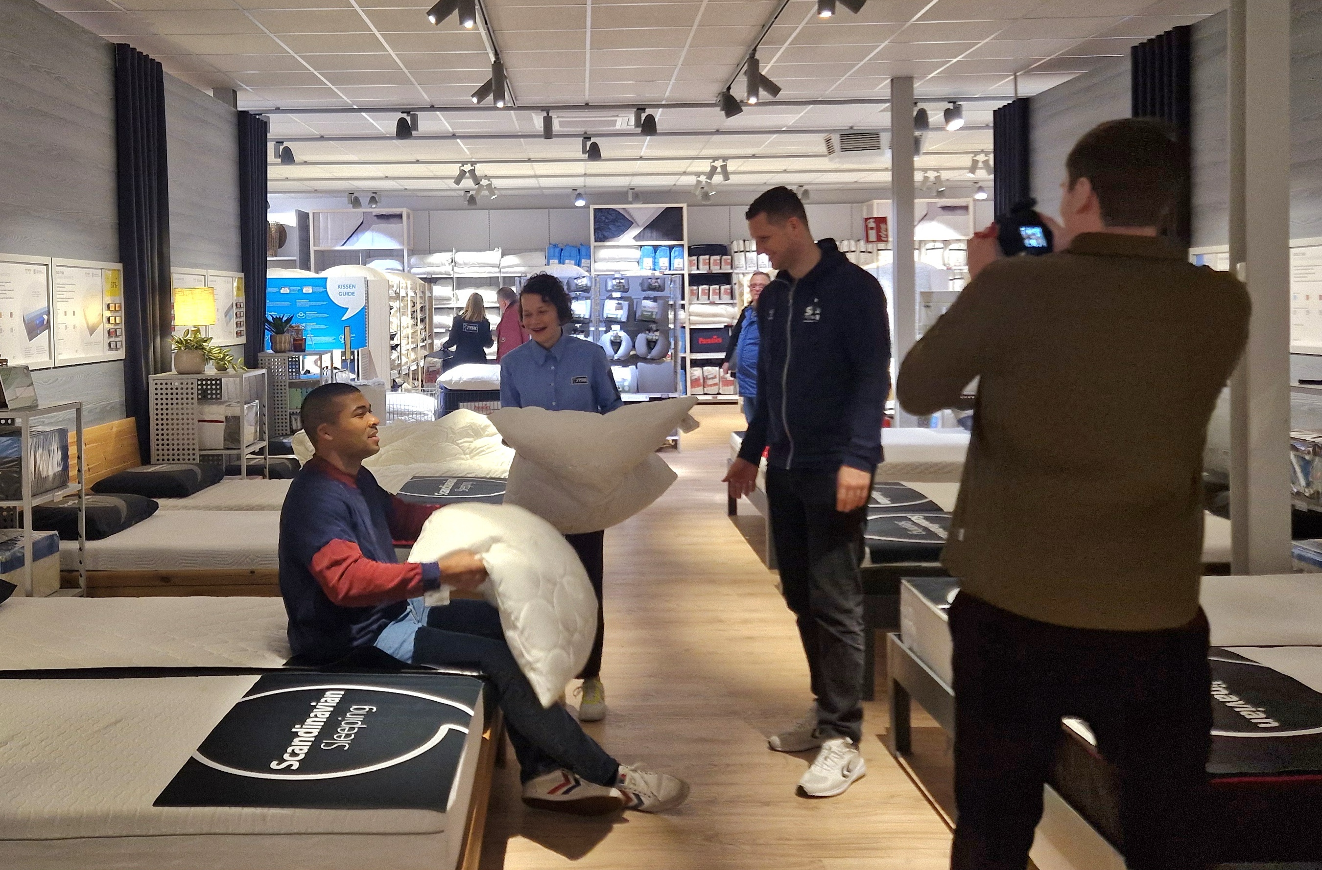 Ein Mann sitzt auf einem Bett im Matratzenstudio. Eine Frau steht daneben und hält ein Kissen in der hand. Ein zweiter Mann steht daneben. Ein dritter Mann filmt alles mit einer Kamera