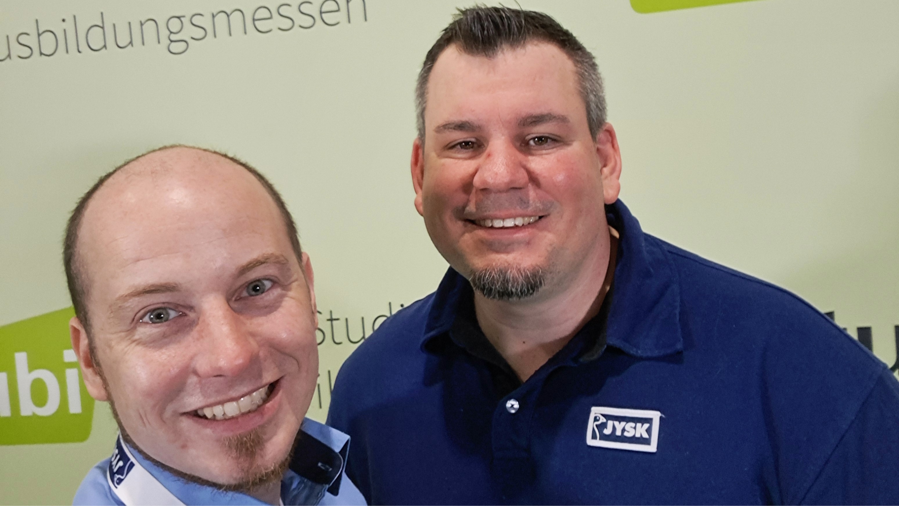 Zwei Männer in JYSK-Arbeitskleidung machen ein Selfie und lachen in die Kamera