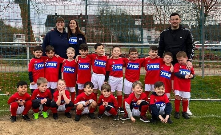 Kleine Jungen, etwa vier bis fünf Jahre alt, sitzen und stehen auf einem Fußballplatz. Sie tragen JYSK-Trikots.