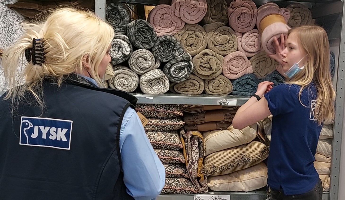 Zwei junge Frauen stehen vor einem Regal mit vielen Decken und unterhalten sich 