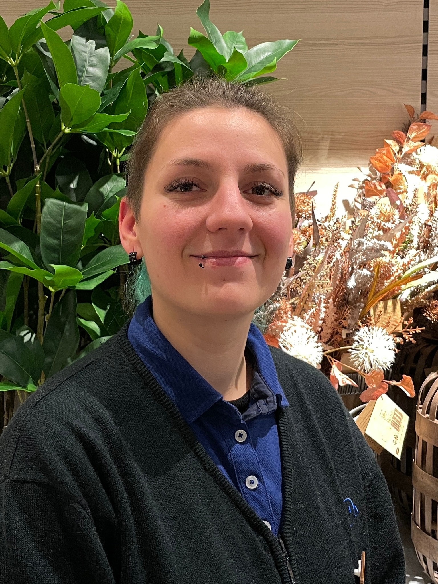 Eine junge Frau lächelt freundlich selbstbewusst in die Kamera. Die junge Frau trägt die Haare zu einem Pferdeschwanz. Sie hat ein Piercing in der Unterlippe. Darüber hinaus trägt sie ein Poloshirt und darüber eine Strickjacke