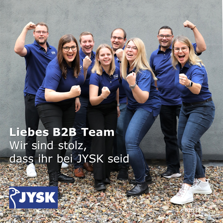 Acht Menschen stehen in zwei Reihen hintereinander, vorne vier Frauen, hinten vier Männer. Sie tragen Poloshirts mit dem JYSK-Logo und lächeln in die Kamera. 