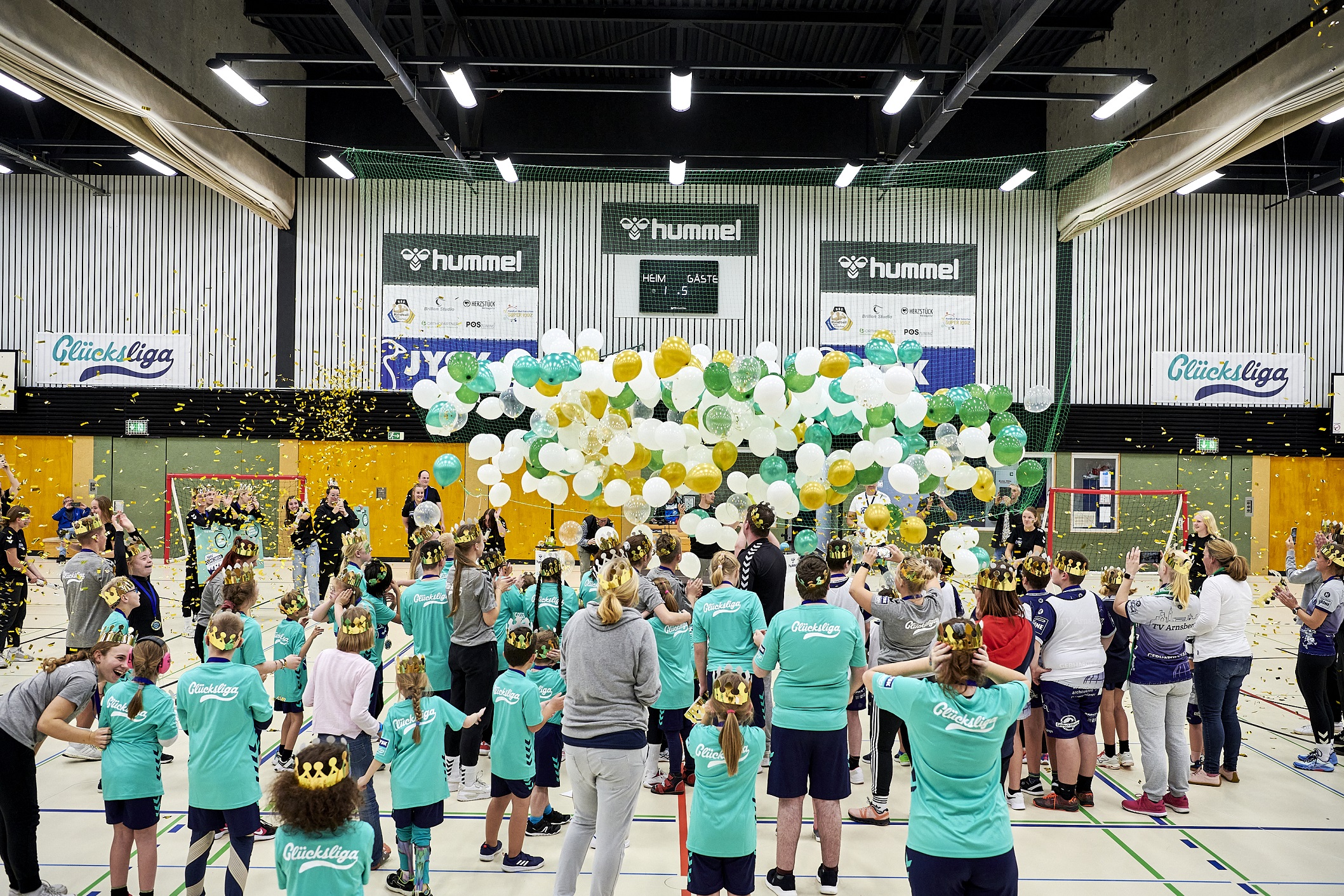Viele Menschen, zumeist Kinder stehen in einer Sporthalle, vor und über ihren Köpfen schweben Luftballons, viele der Menschen tragen eine Krone 