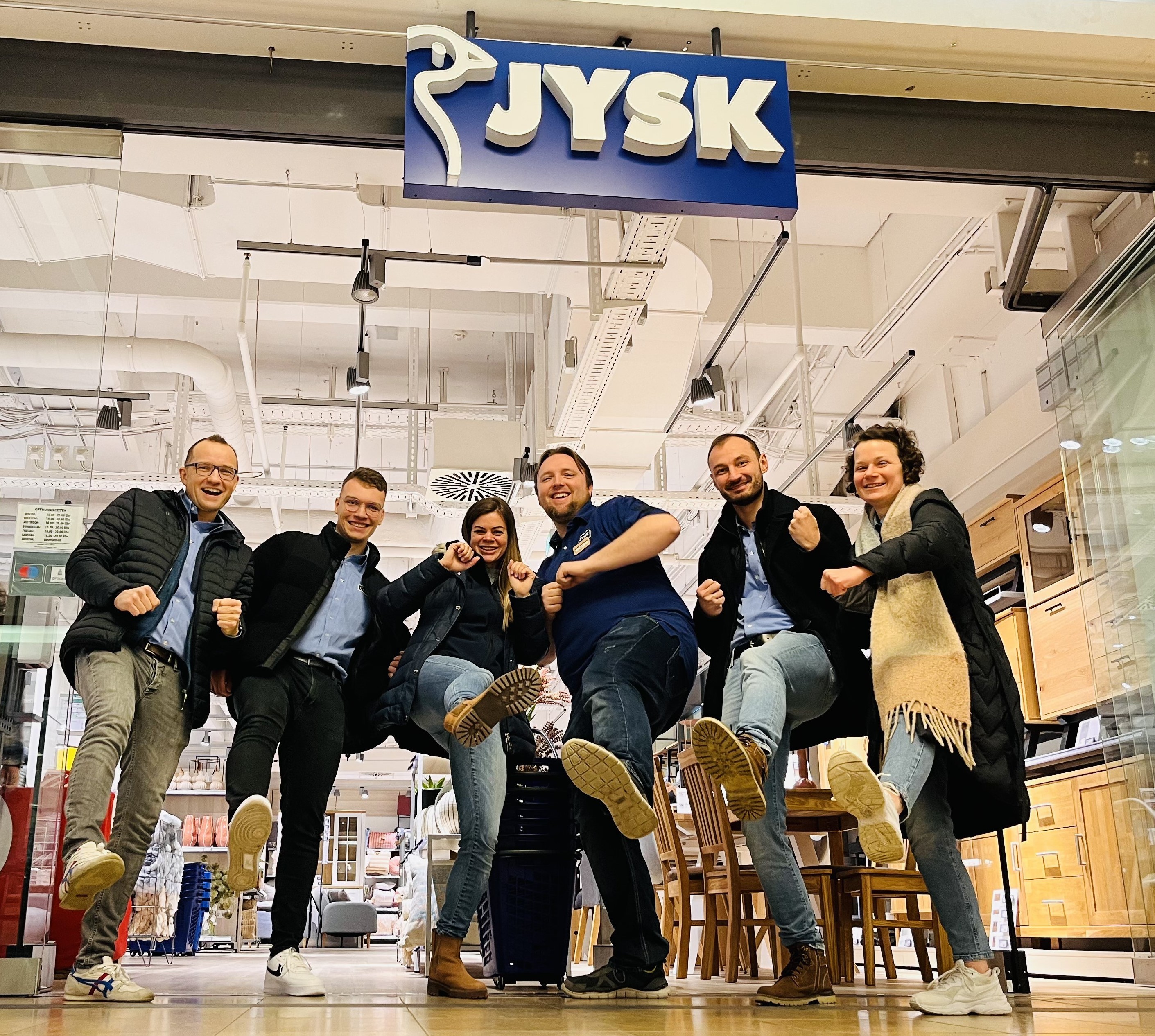 Sechs Personen – zwei Frauen und vier Männer – lächeln in eine Kamera, die auf dem Fußboden platziert ist. Sie machen eine Kick-Bewegung mit ihren Beinen. Über ihnen ist das JYSK-logo zu sehen