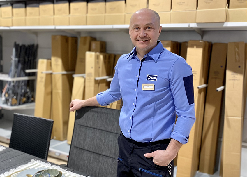Denis står vid utemöblerna. Bakom honom är det förpackningar i en hylla.