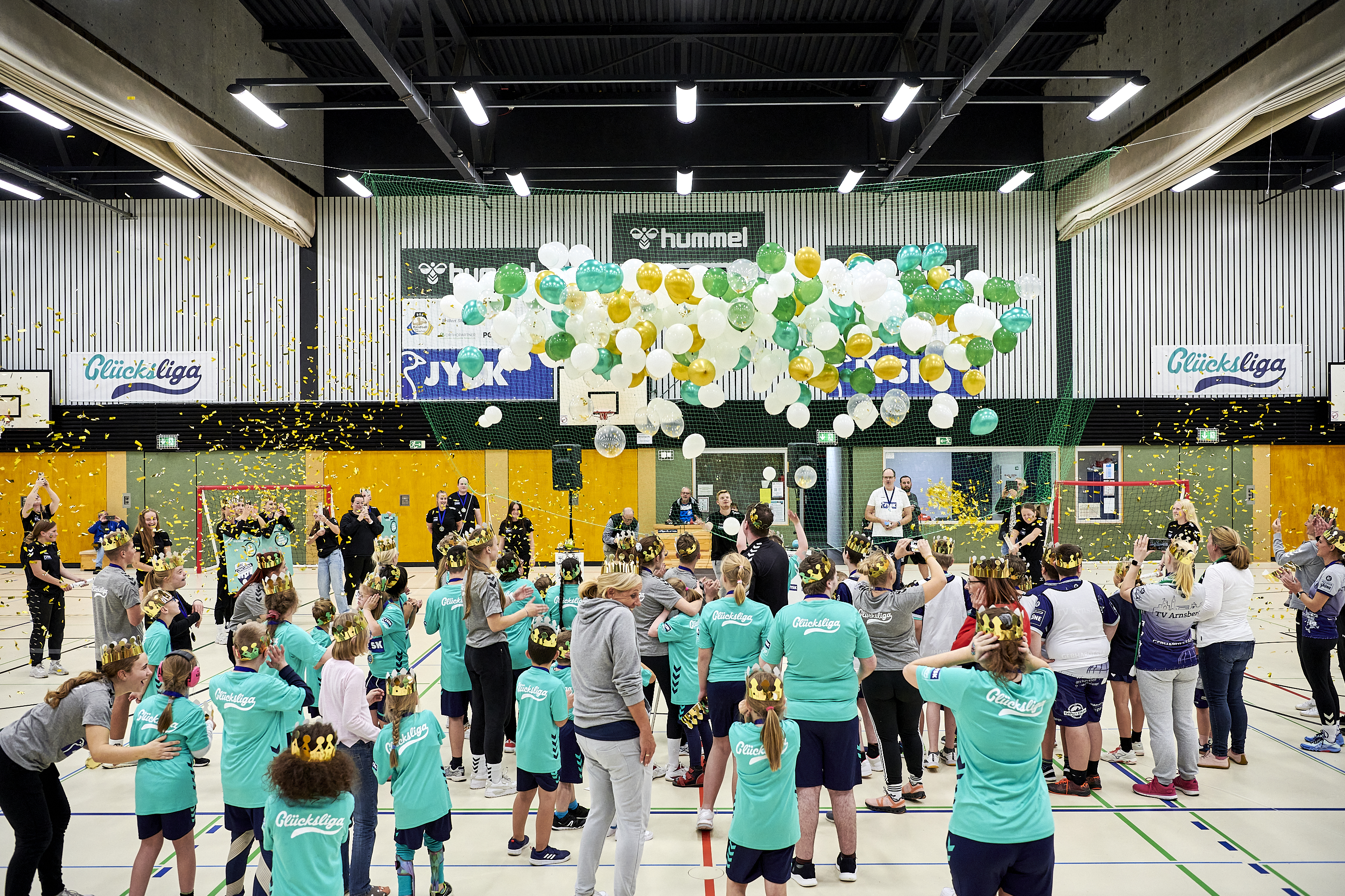 Eine große Gruppe an Menschen, die meisten von ihnen Kinder, stehen in einer Turnhalle. Über ihnen fallen Luftballons von der Hallendecke