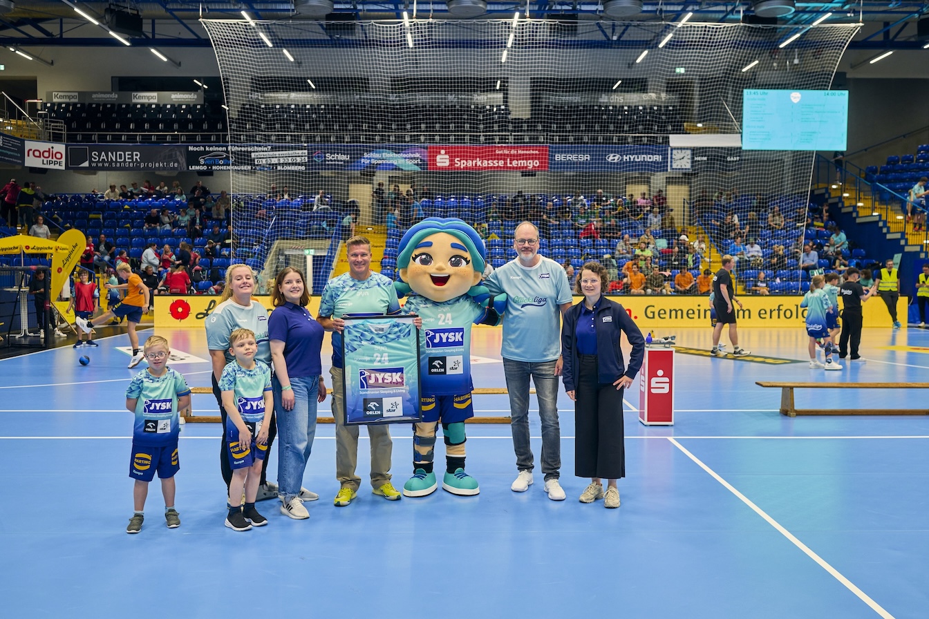 Sieben Menschen und ein Maskottchen stehen in einer Handballhalle