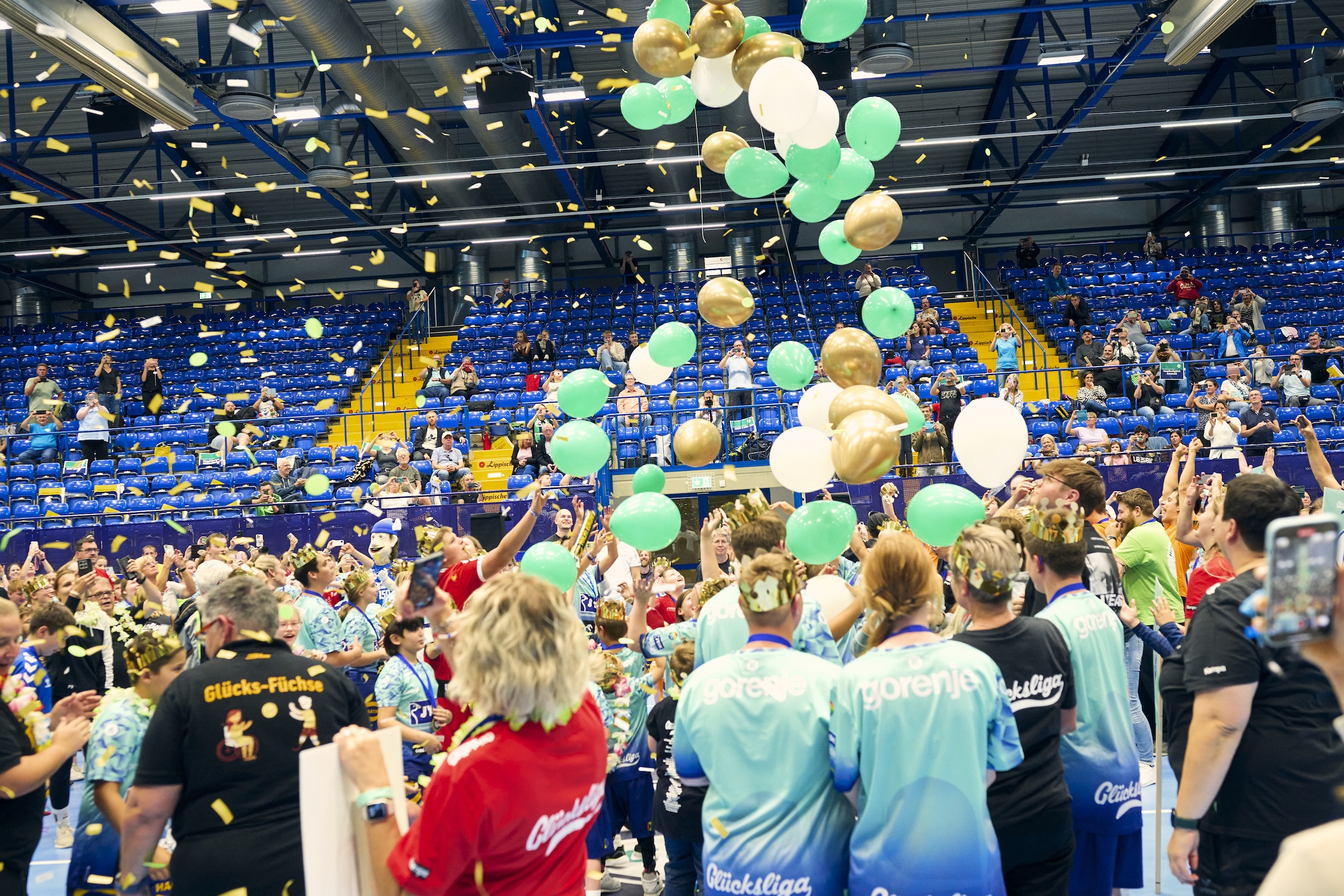 Viele Kinder stehen mit Kronen auf den Köpfen in einer Turnhalle. Von der Decke regnet es Luftballons