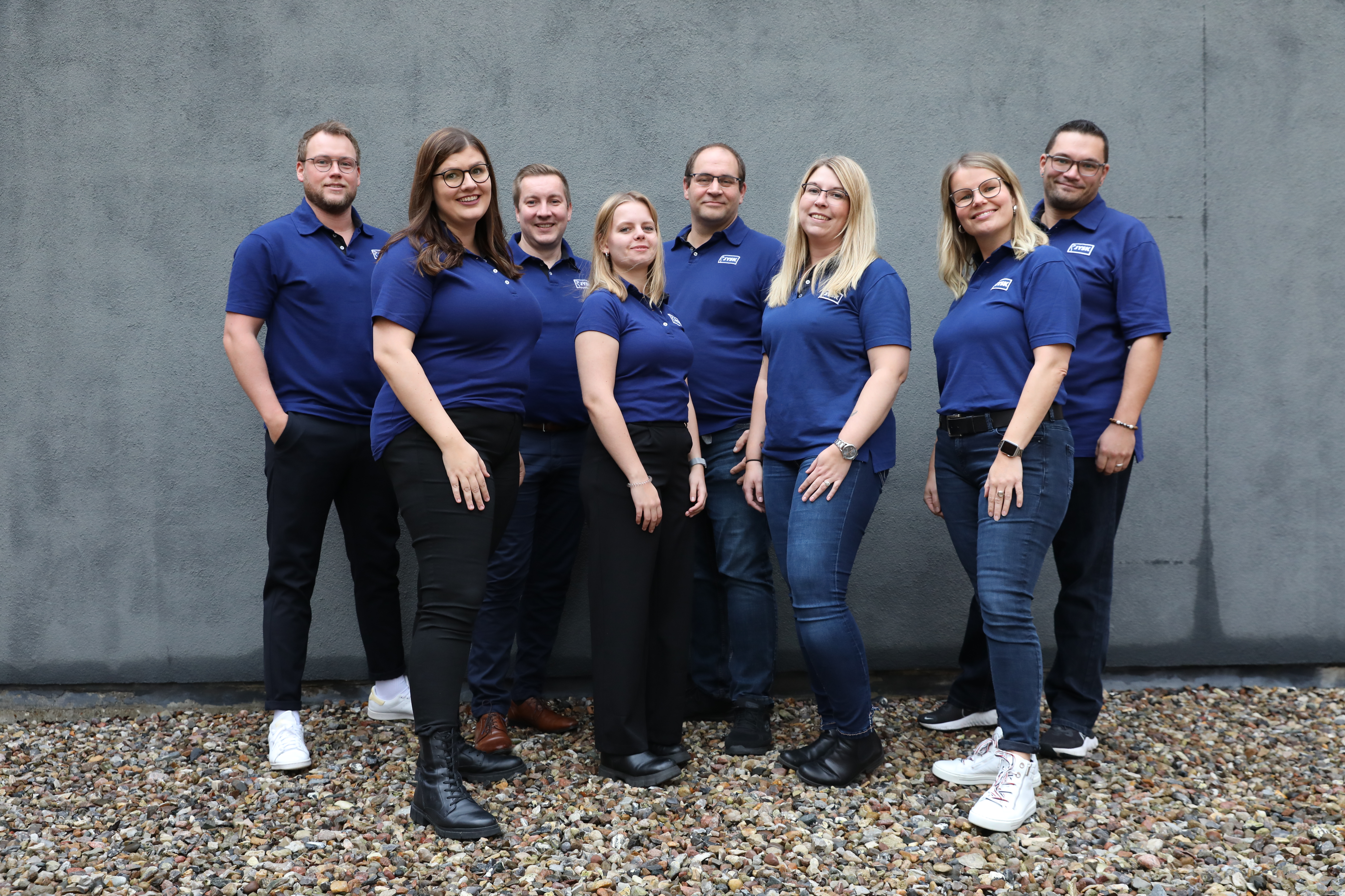 Acht Menschen stehen in zwei Reihen hintereinander, vorne vier Frauen, hinten vier Männer. Sie tragen Poloshirts mit dem JYSK-Logo und lächeln in die Kamera. 