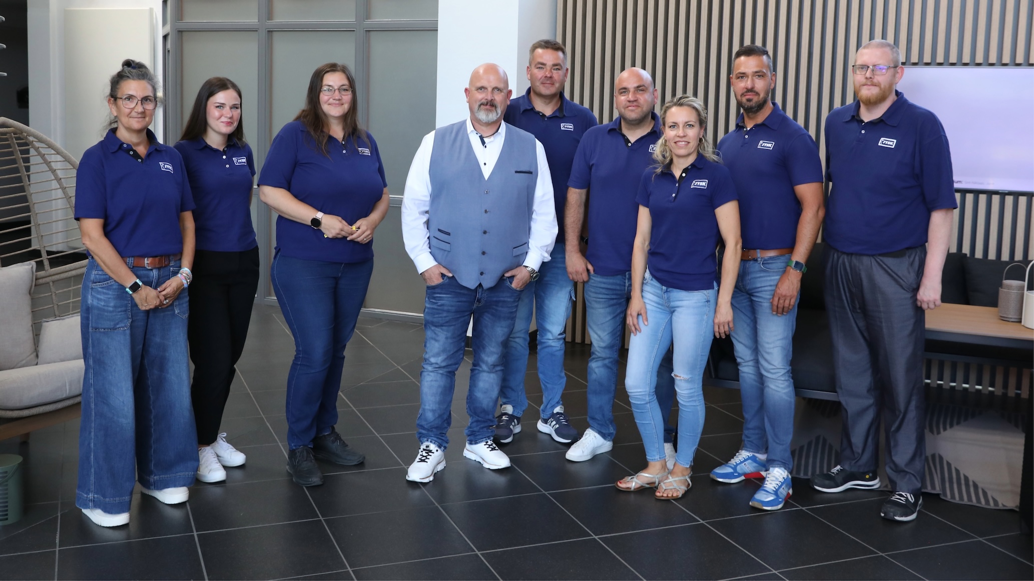 Neun Menschen – drei Frauen und sechs Männer – stehen im Foyer der JYSK Deutschland-Zentrale. Die meisten von ihnen tragen JYSK-Poloshirts. Alle lächeln in die Kamera