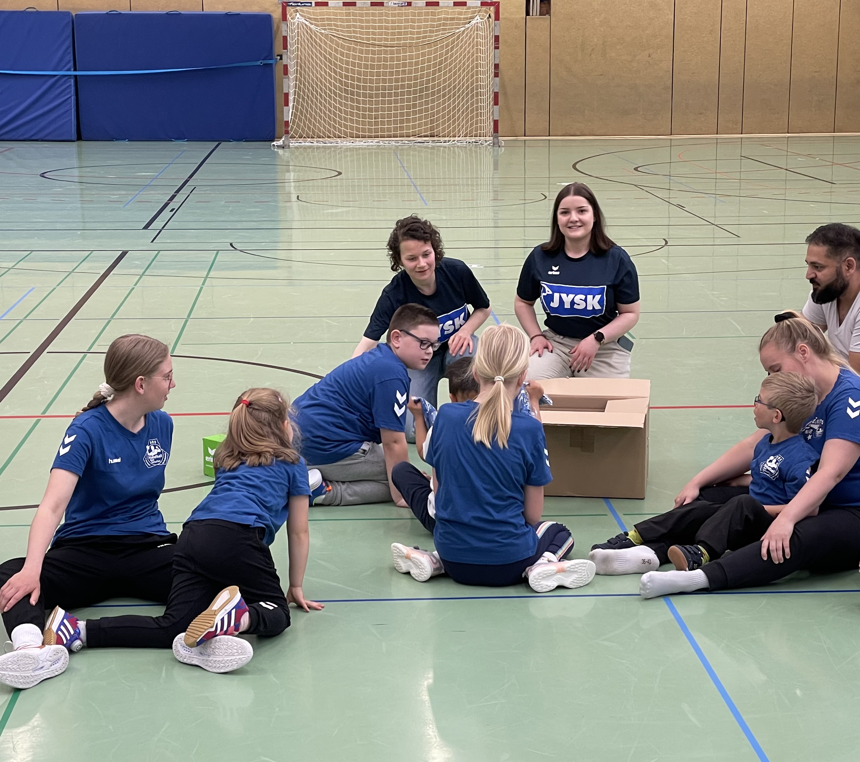 Zwei junge Frauen hocken hinter einem Karton, aus dem sie Trikots nehmen. Mehrere Kinder schauen sie ganz gespannt an.