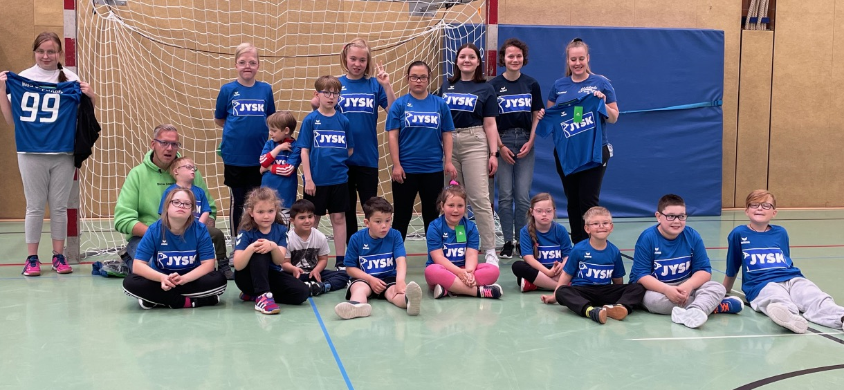 Viele Menschen, zumeist Kinder zwischen 5 und 12 Jahren stehen und sitzen vor einem Handballtor. Sie strahlen in die Kamera, weil sie sich über ihre neuen Trikots freuen.