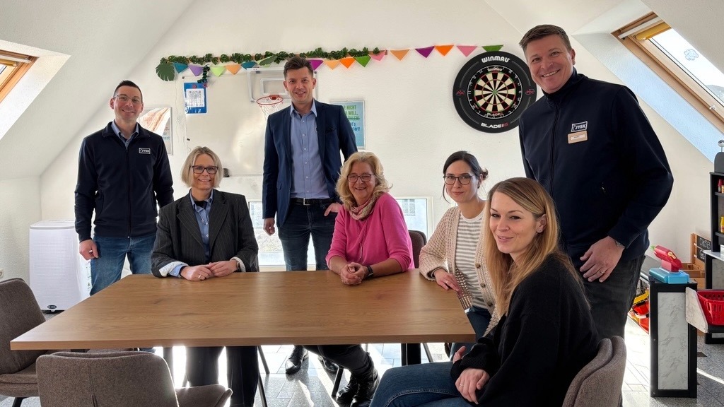Mehrere Menschen sitzen und stehen um einen großen Tisch herum und lächeln in die Kamera