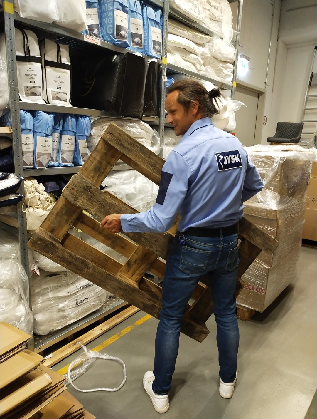 Ein Mann mit Dutt steht in einem Lager und hält eine Palette in den Händen. Er trägt ein JYSK-Hemd