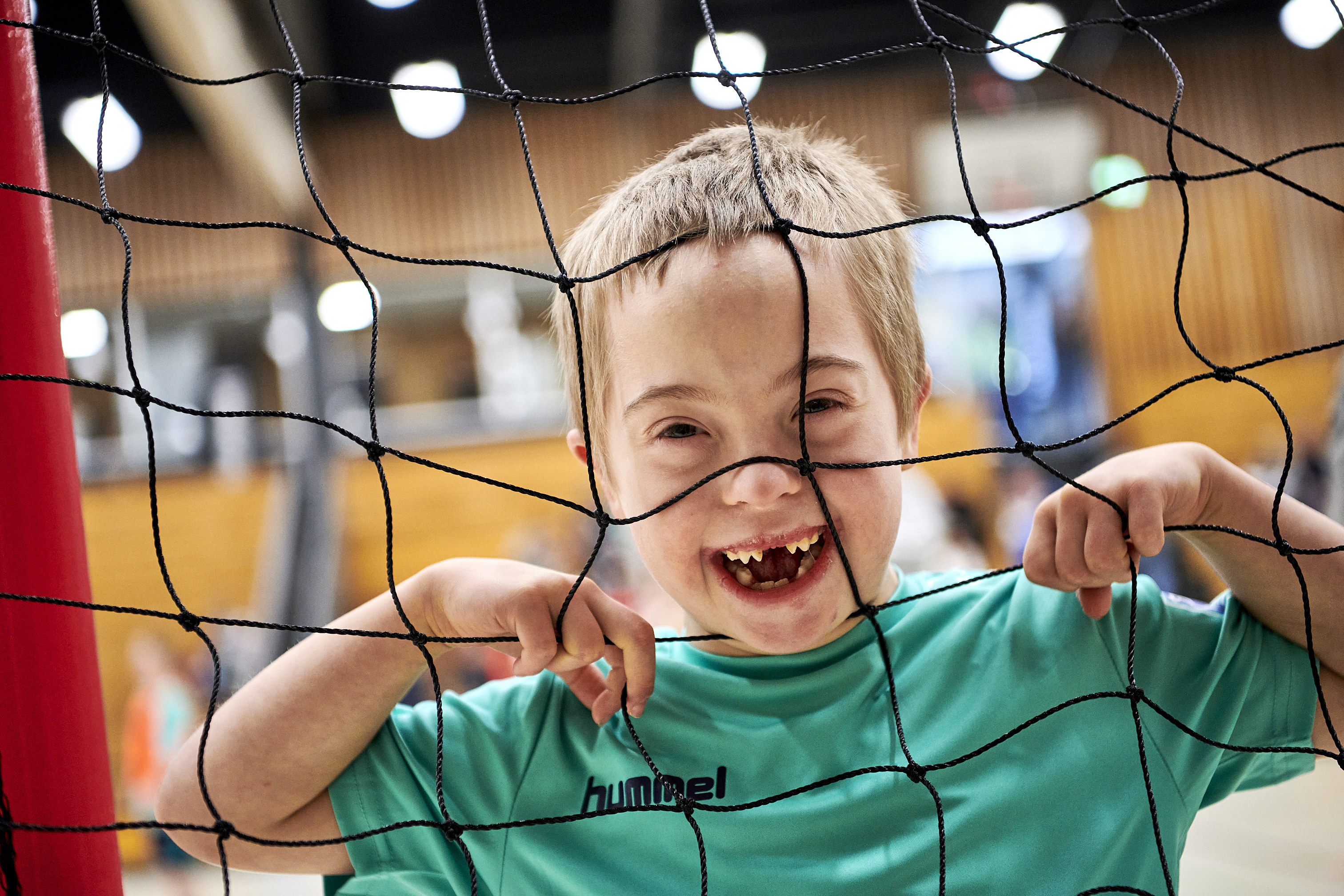 Ein Junge mit kurzen Haaren steht in einem Handballtor und schaut durch das Netz in die Kamera. Er lacht ganz doll und zeigt eine große Zahnlücke