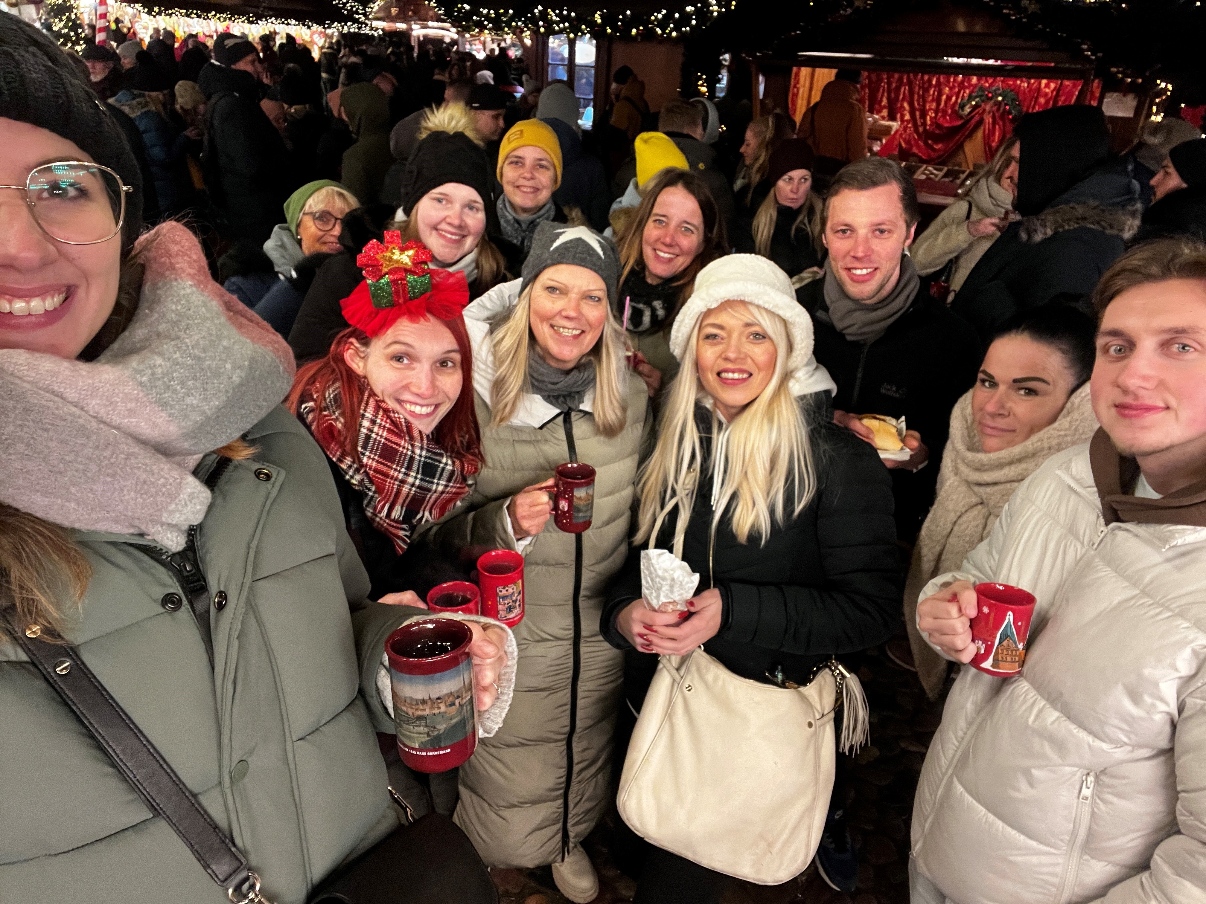 Eine größere Gruppe steht auf einem Weihnachtsmarkt. Eine junge Frau am linken Bildrand macht ein Selfie der Gruppe.