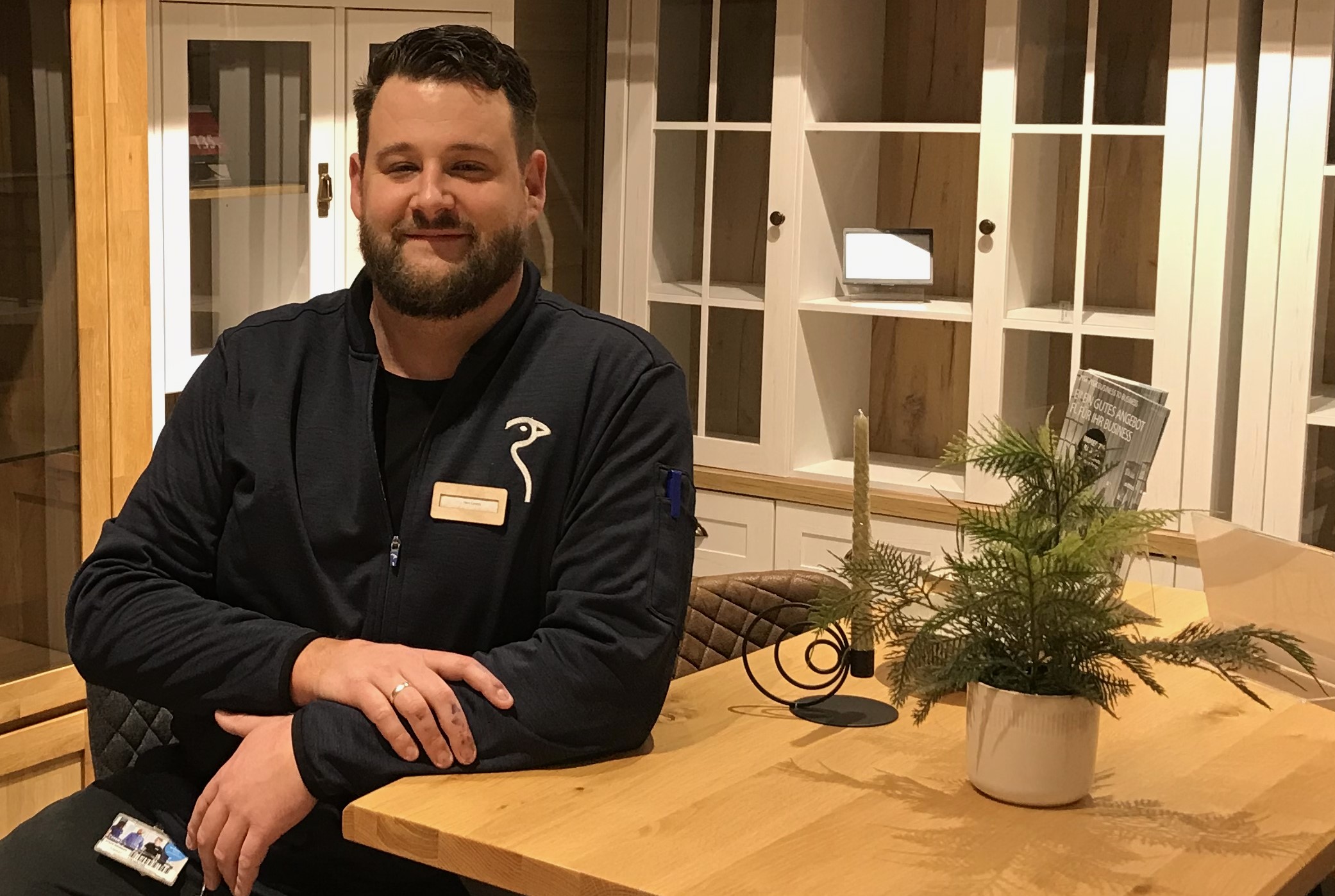 Ein Mann Mitte 30 sitzt an einem Tisch, auf dem eine Pflanze steht. Der Mann trägt einen Bart und eine Jacke mit JYSK-Logo. Er lächelt in die Kamera