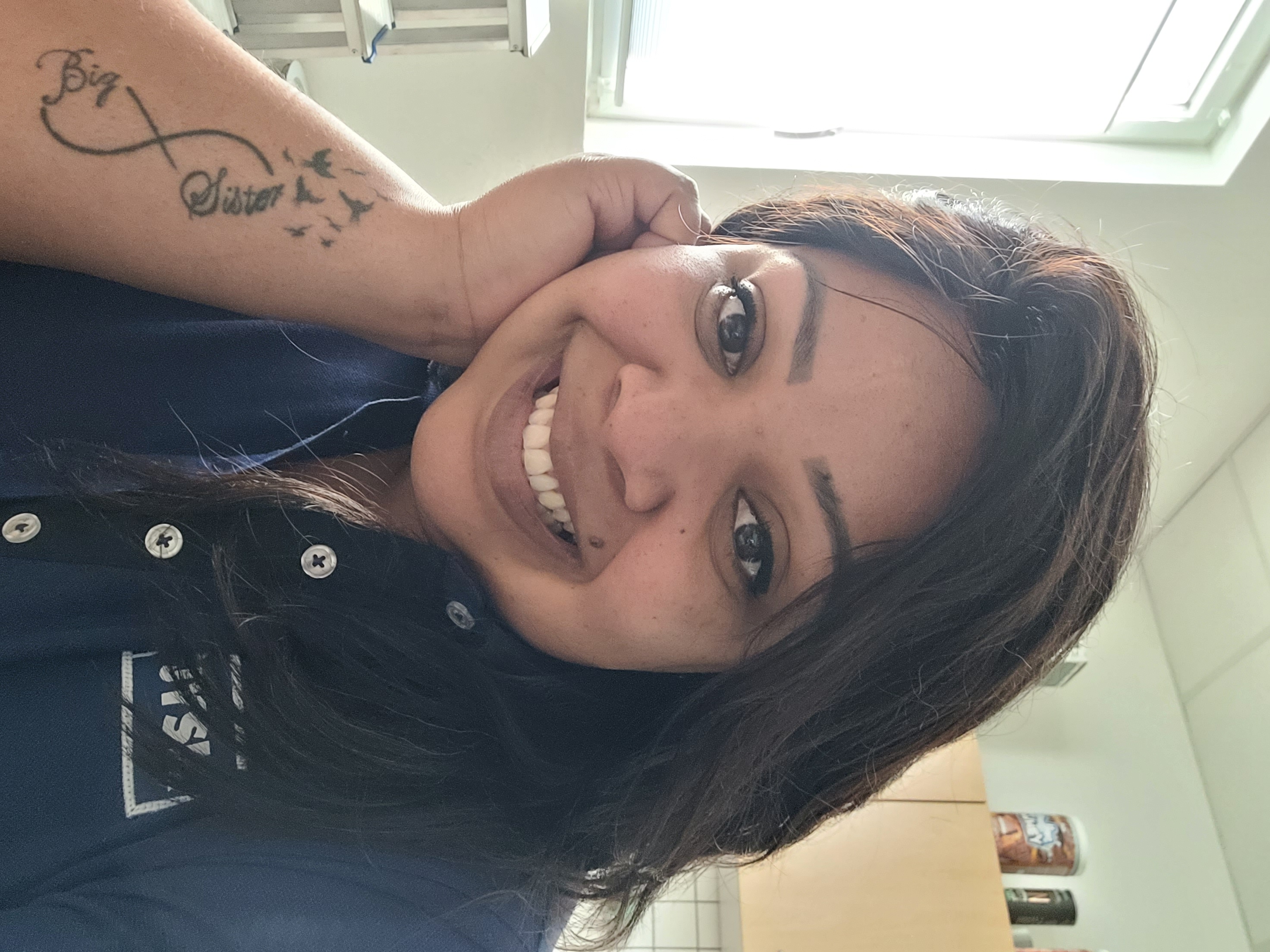 Eine junge Frau mit langen Haaren lächelt in eine Kamera. Sie trägt ein JYSK_Poloshirt, am rechten Arm trägt sie ein Tattoo