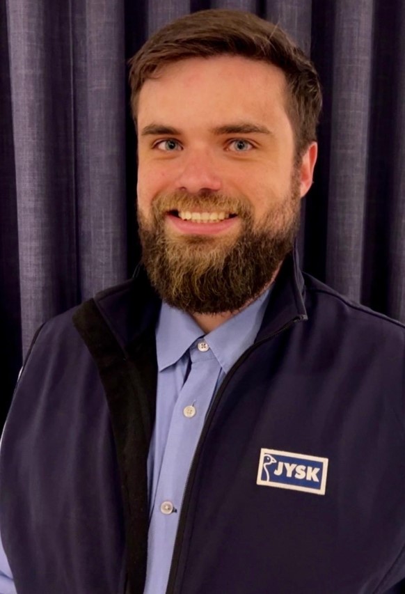 Ein junger Mann mit Dreitagebart, JYSK-Hemd und JYSK-Poloshirt lächelt in die Kamera