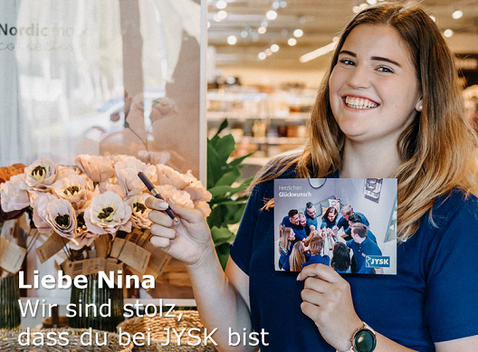 Eine junge Frau mit langen Haaren grinst fröhlich in eine Kamera, sie hält einen Stift in der Hand.