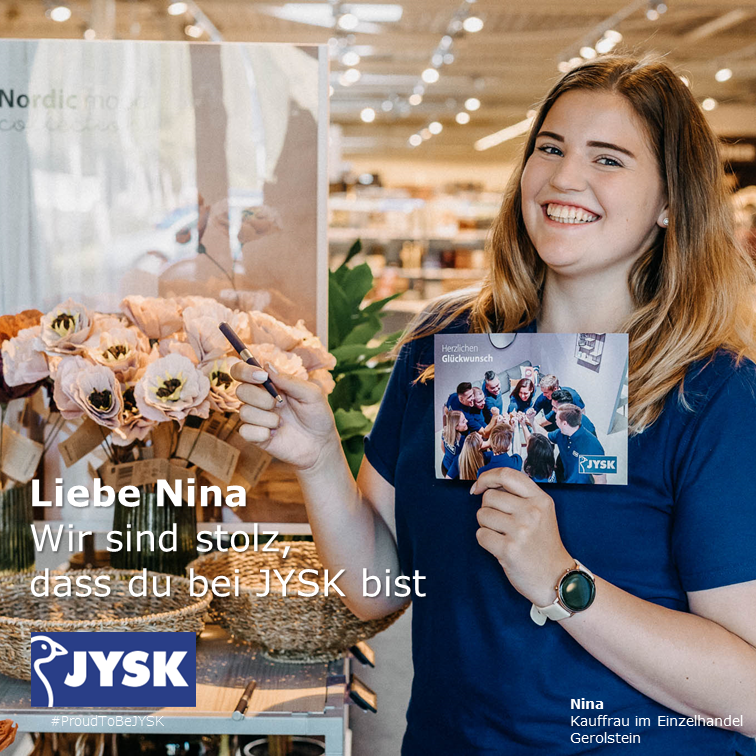 Eine junge Frau mit längeren Haaren hält einen Stift und eine Karte in der Hand. Sie lächelt selbstbewusst in die Kamera. Sie steht in einem JYSK Store