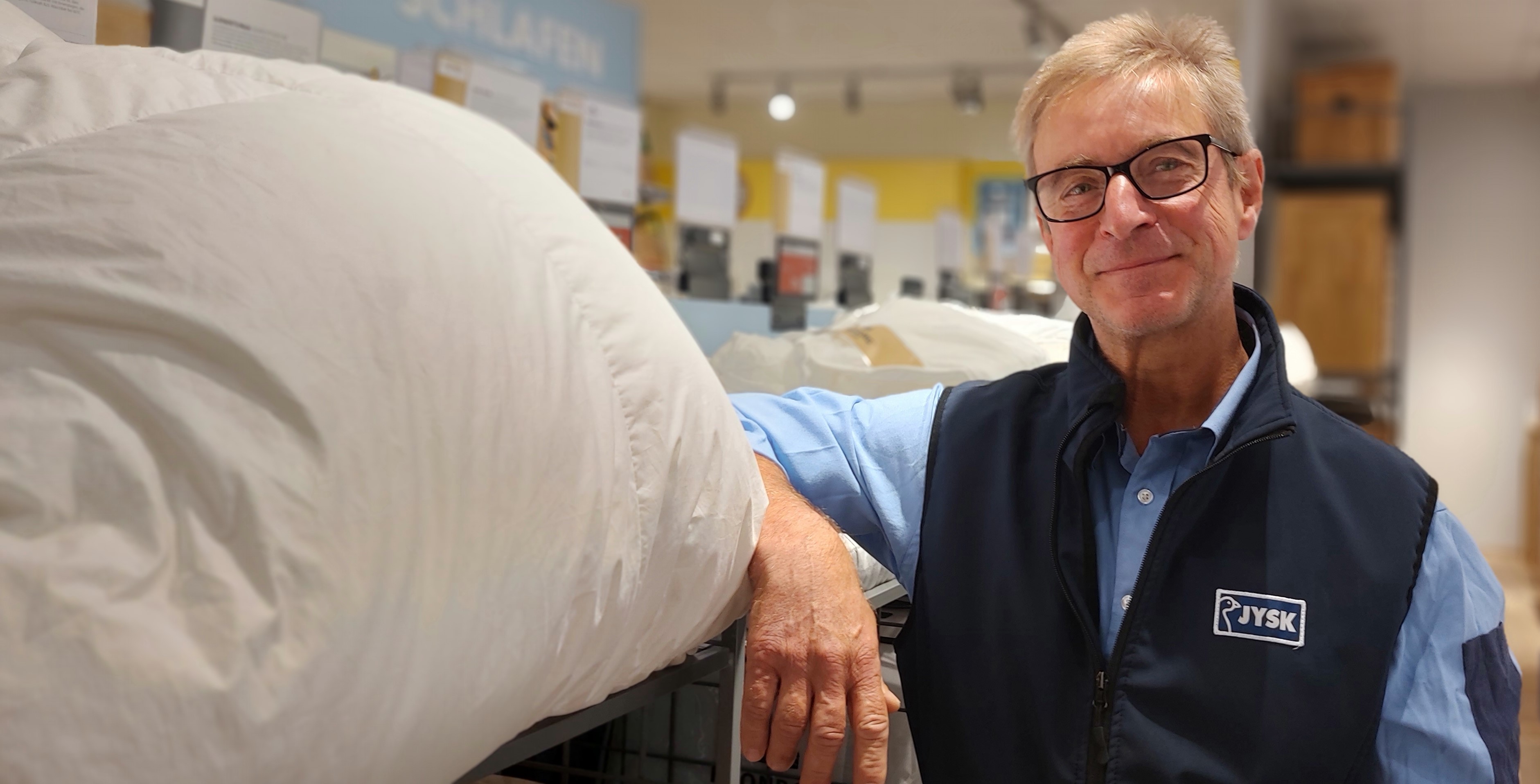 Ein Mann mit Brille und kurzen Haaren lächelt in die Kamera. Er lehnt lässig an einem Regal, aus dem ein Kissen rausschaut.