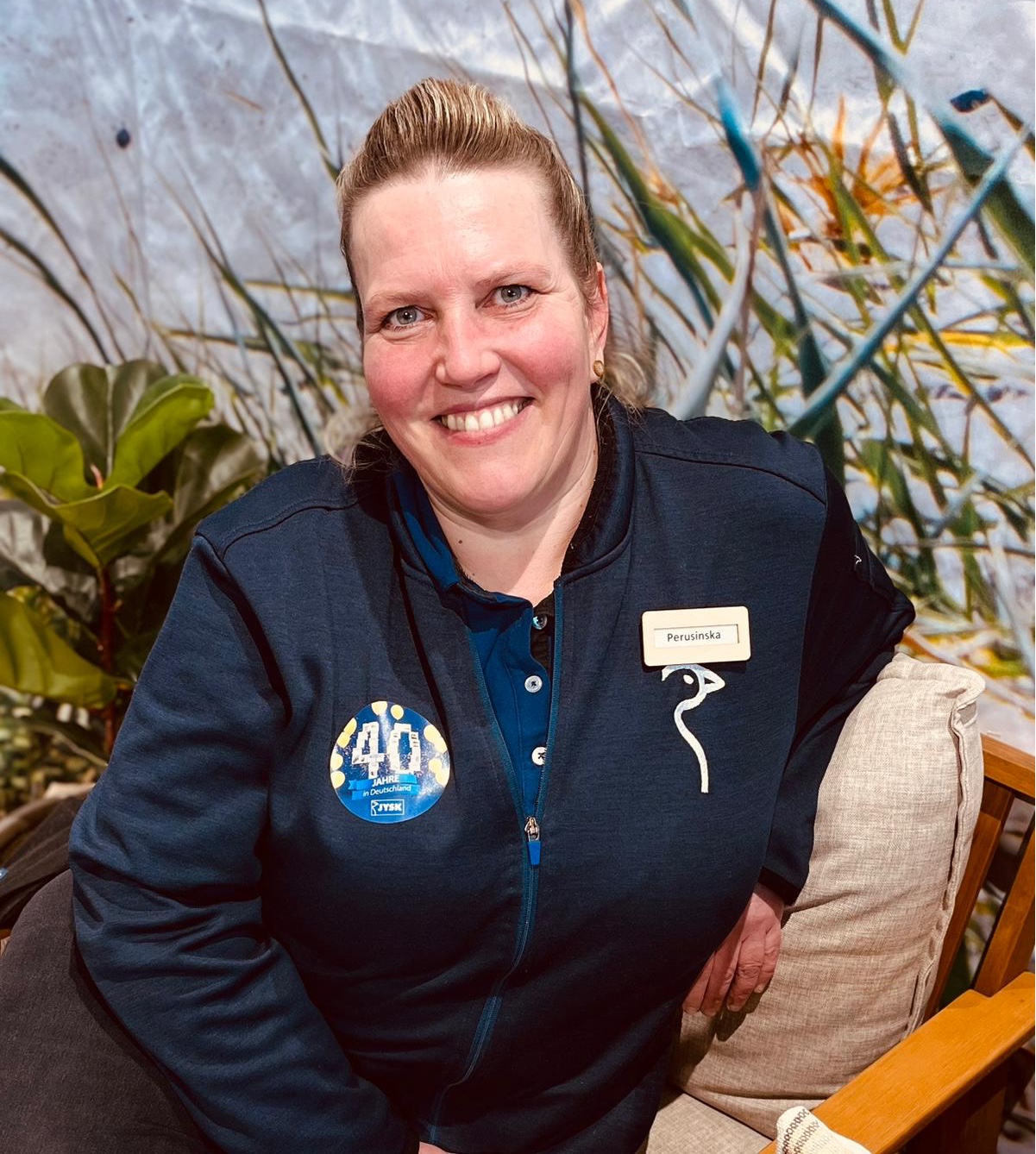 Eine Frau mit Pferdeschwanz sitzt auf einem Sofa und lächelt in die Kamera. Sie trägt ein JYSK_Poloshirt und darüber eine JYSK_Strickjacke