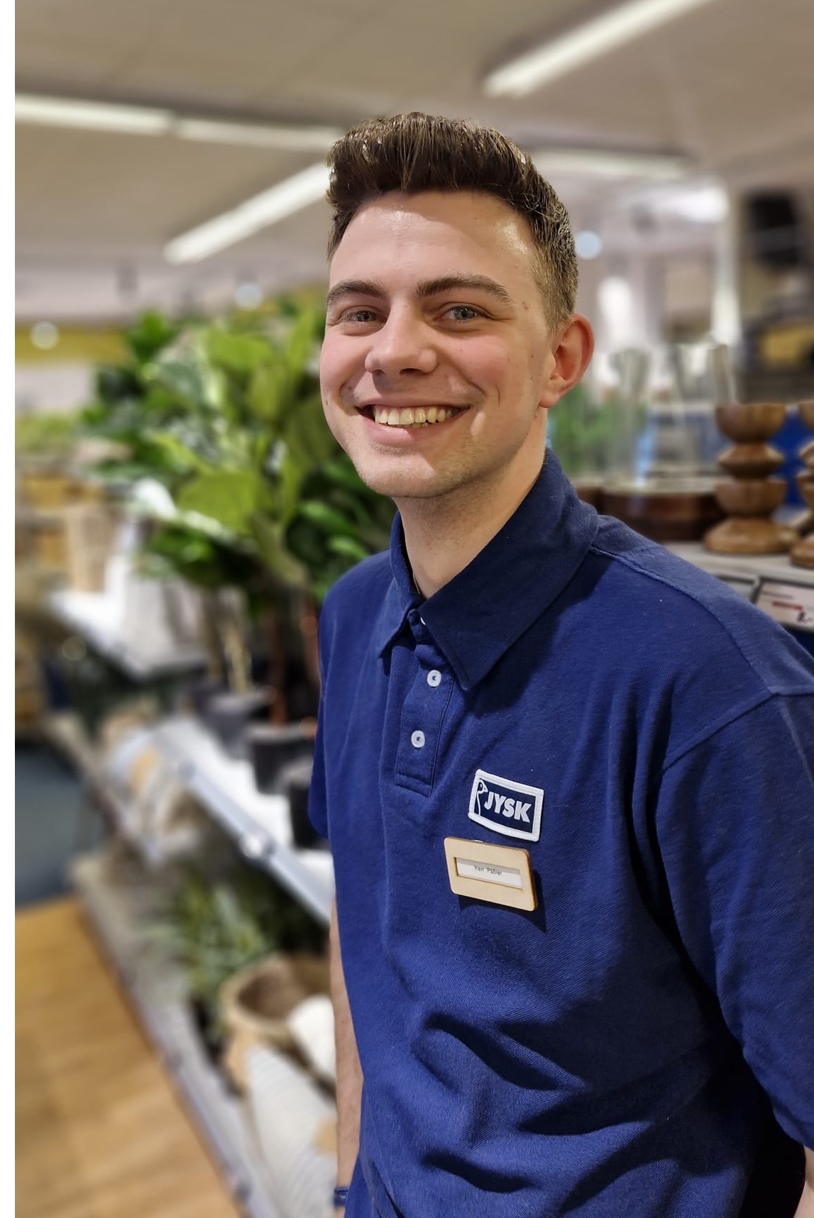 Ein junger Mann im JYSK_Poloshirt lächelt freundlich in die Kamera. Im Hintergrund ist der Innenraum eines JYSK Stores zu erkennen