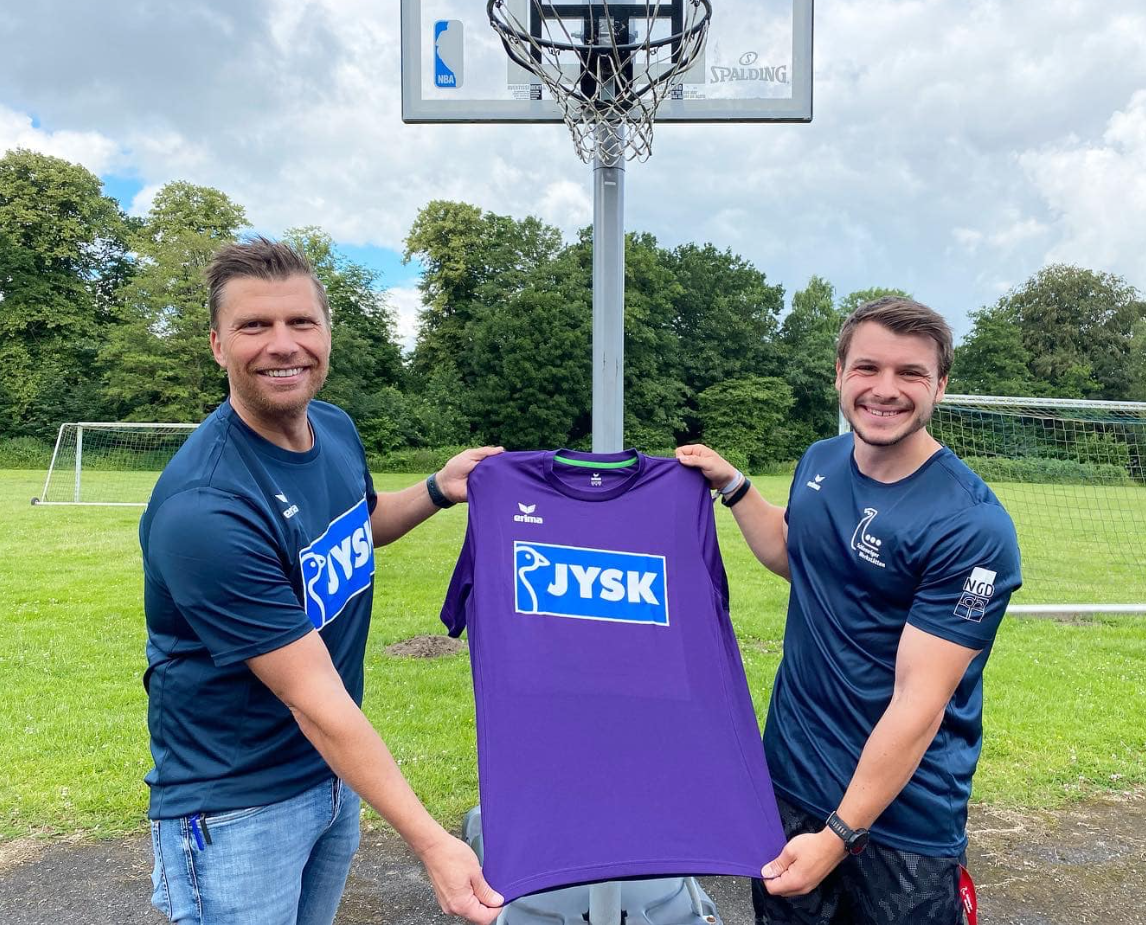 Zwei Männer halten gemeinsam ein Trikot in den Händen und lächeln. Im Hintergrund ist ein Basketballkorb zu sehen