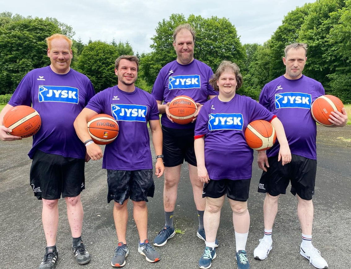 Fünf Menschen in Trikots und kurzen Hosen stehen mit Basketbällen in den Händen auf einem Schotterplatz und lächeln