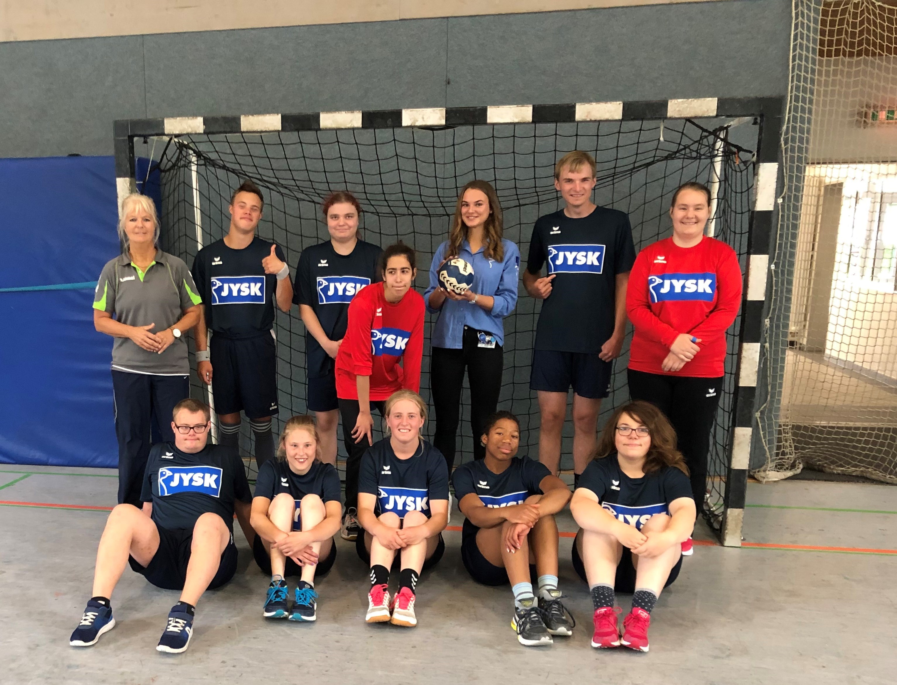 Einige Kinder stehen in einem Handballtor, eine junge Frau hält einen Handball in der Hand