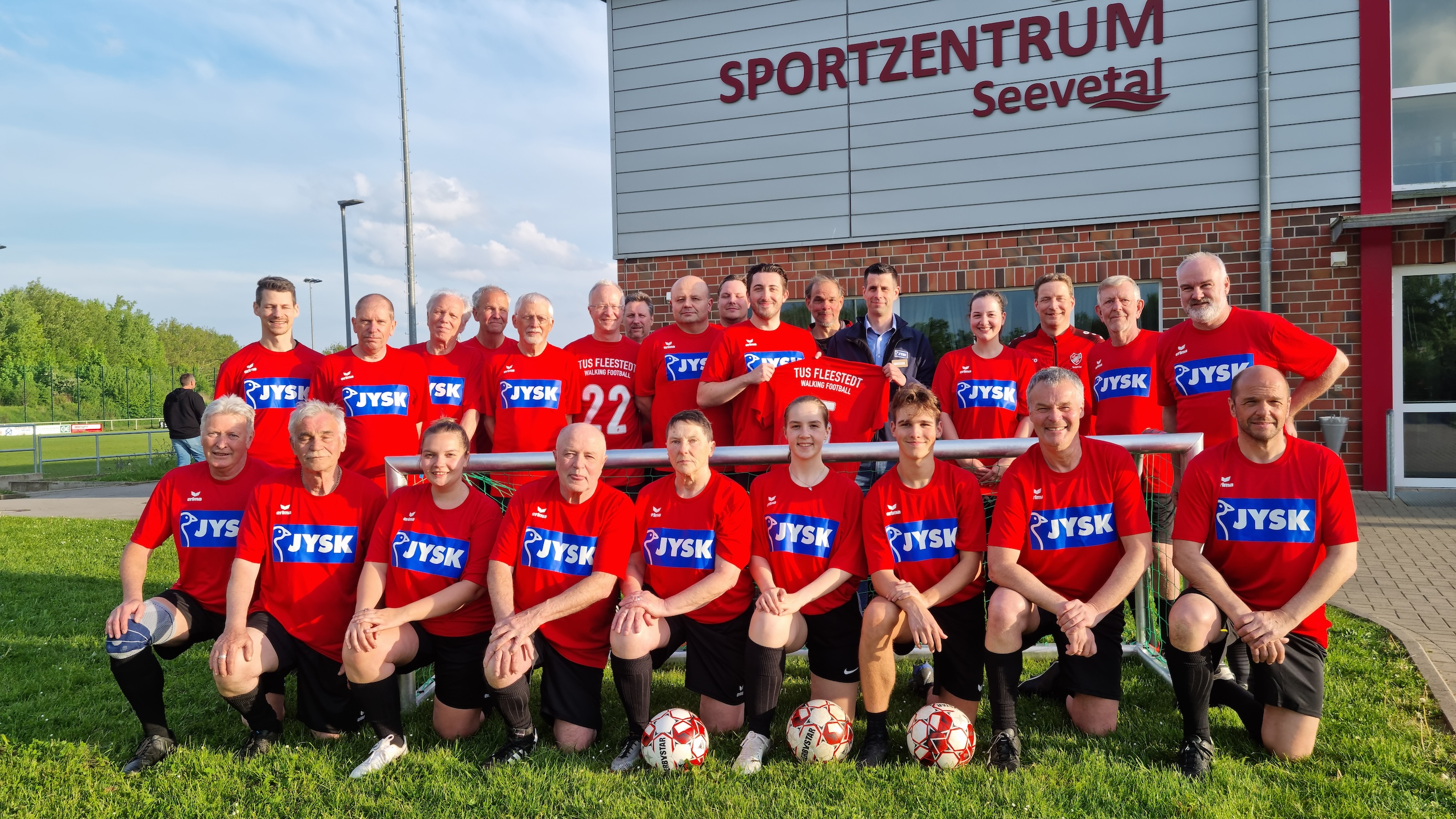 Viele Menschen – sowohl jung als auch alt – stehen und hocken auf einem Fußballplatz. Sie lächeln in die Kamera. Im Hintergrund ist ein Clubhaus zu erkennen.