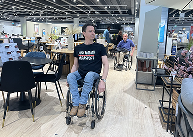 Två män ser sig om i en butik. De befinner sig bland bord och stolar. I förgrunden syns Henrik Larsson från FIFH. Han siter i en rullstol och har ena handen på hjulet som i framåtrörelse. Han har på sig en svart tröja där det står FIFH Malmö Parasport och ett par blåa jeans. Han tittar åt vänster. Bakom honom syns Peter Nilsson, också han är på väg framåt i sin rullstol. Han har på sig en blå skjorta och ett par beigea byxor. Han har blicken framåtvänd. I bakgrunden står två kunder.