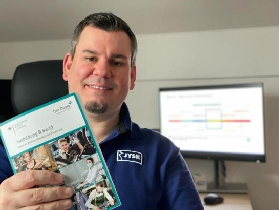 Ein Mann mit Kinnbart ung JYSK-Poloshirt hält ein Heftchen mit der Aufschrift "Ausbildung & Beruf" in der Hand. Im Hintergrund ist leicht verschwommen ein Computerbildschirm zu erkennen. 