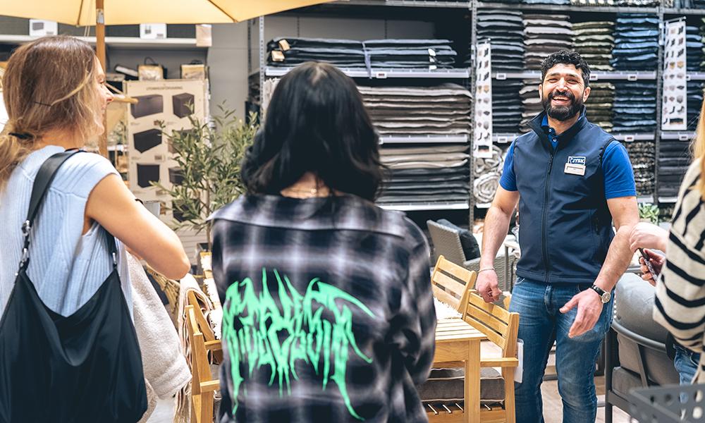 Influencers som lyssnar till en JYSK medarbetare. Medarbetaren sstår framför en ställning som är full med stolsdynor. Han skrattar.