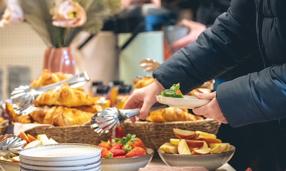 Händer plockar från ett fikabord dukat med croissanter, smothies och jordgubbar.