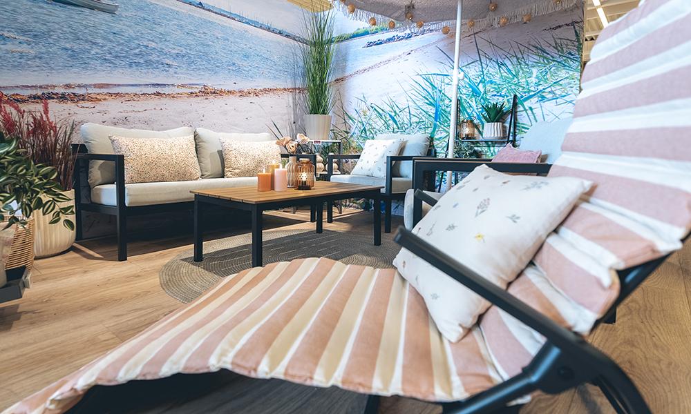 En solstol med en randig dyna i förgrunden. Bakom står ett svart loungeset mot en bakgrund som påminner om en sandstrand. 