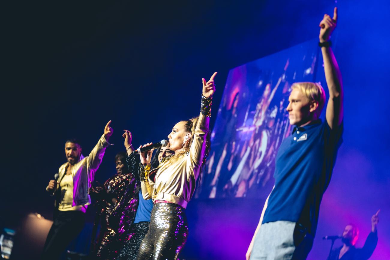 Två JYSK-medarbetare dansar tillsammans med Lina Hedlund på scen. En grupp JYSK-medarbetare dansar på scenen mitt i lokalen. Foto: Joakim Thörne.