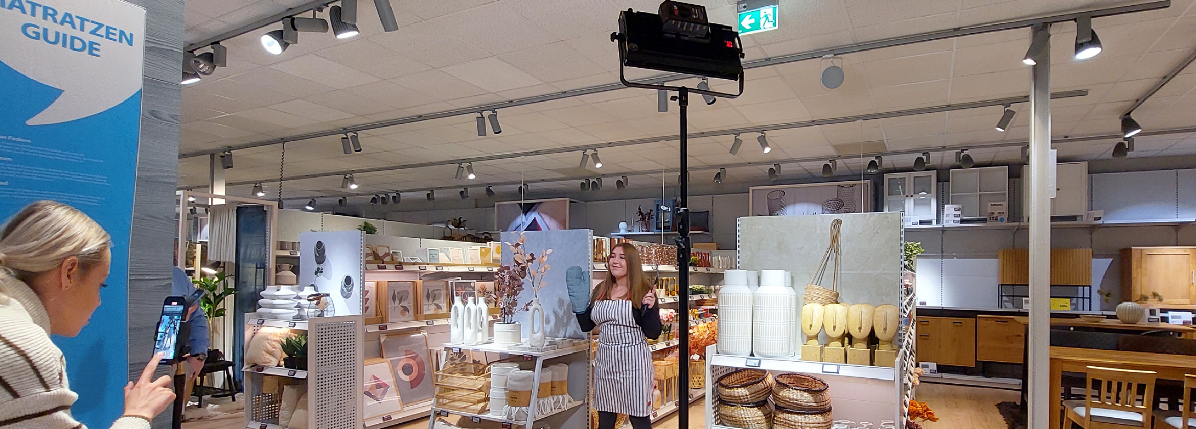 Eine junge Frau mit langen Haaren steht in einem Store. Sie trägt eine Schürze und einen Backhandschuh. Eine weitere Frau steht hinter einem Handy und nimmt die Szene auf. 