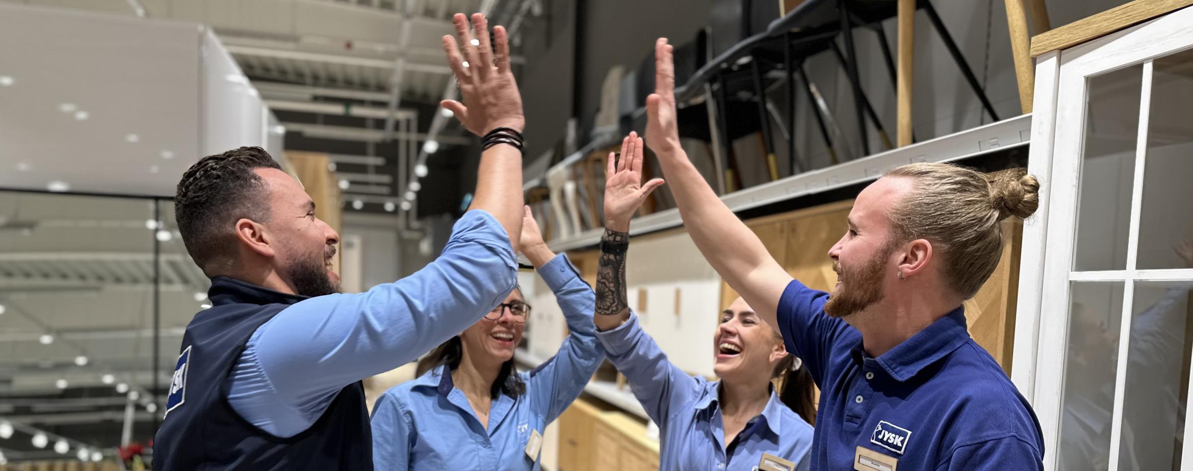 Fyra butikschefstraineer gör high five i butiksmiljö. 