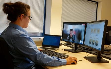 Eine Frau mit Brille sitzt vor einem PC. Auf dem Bildschirm ist ein Mann zu erkennen