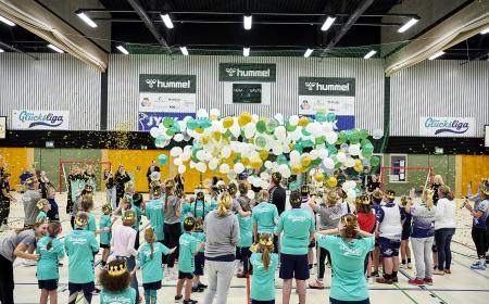 Viele Menschen, zumeist Kinder stehen in einer Sporthalle, vor und über ihren Köpfen schweben Luftballons, viele der Menschen tragen eine Krone 