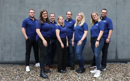 Acht Menschen stehen in zwei Reihen hintereinander, vorne vier Frauen, hinten vier Männer. Sie tragen Poloshirts mit dem JYSK-Logo und lächeln in die Kamera. 